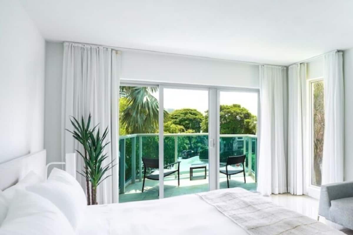 Quarto de hotel moderno com uma cama grande, um sofá cinzento, uma televisão de tela plana e uma varanda com vista para o verde.