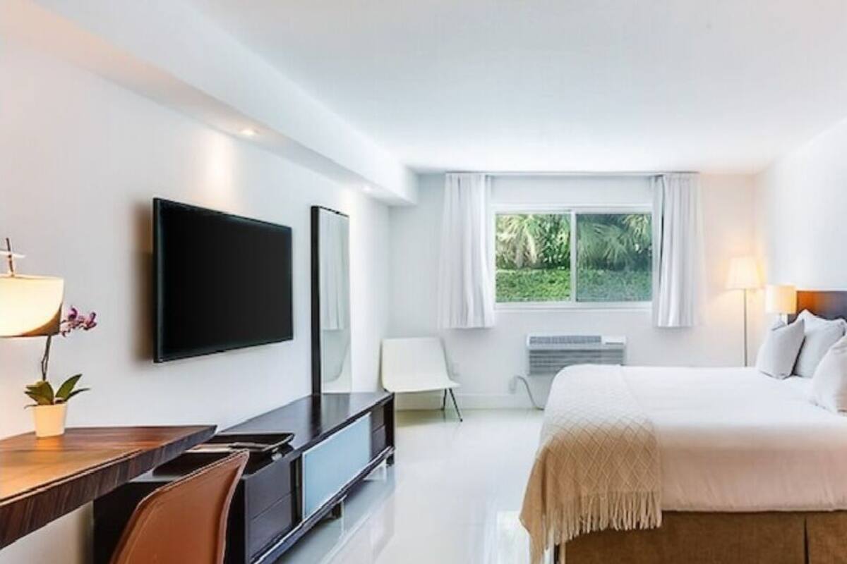 Quarto de hotel moderno com uma cama grande, uma TV de tela plana, uma mesa com cadeira e uma janela com vista para o verde.