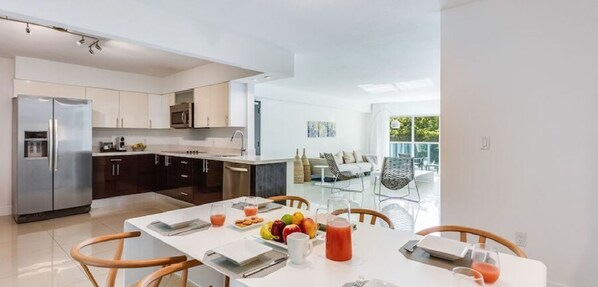 A modern kitchen with a dining area, featuring a refrigerator, microwave, and dining table set with fruit and drinks. - Beach Haus Bal Harbour (Bal Harbour)