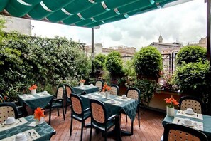 An outdoor patio with tables set for dining, surrounded by lush greenery and a view of buildings in the background. - Condominio Monti (Rome)