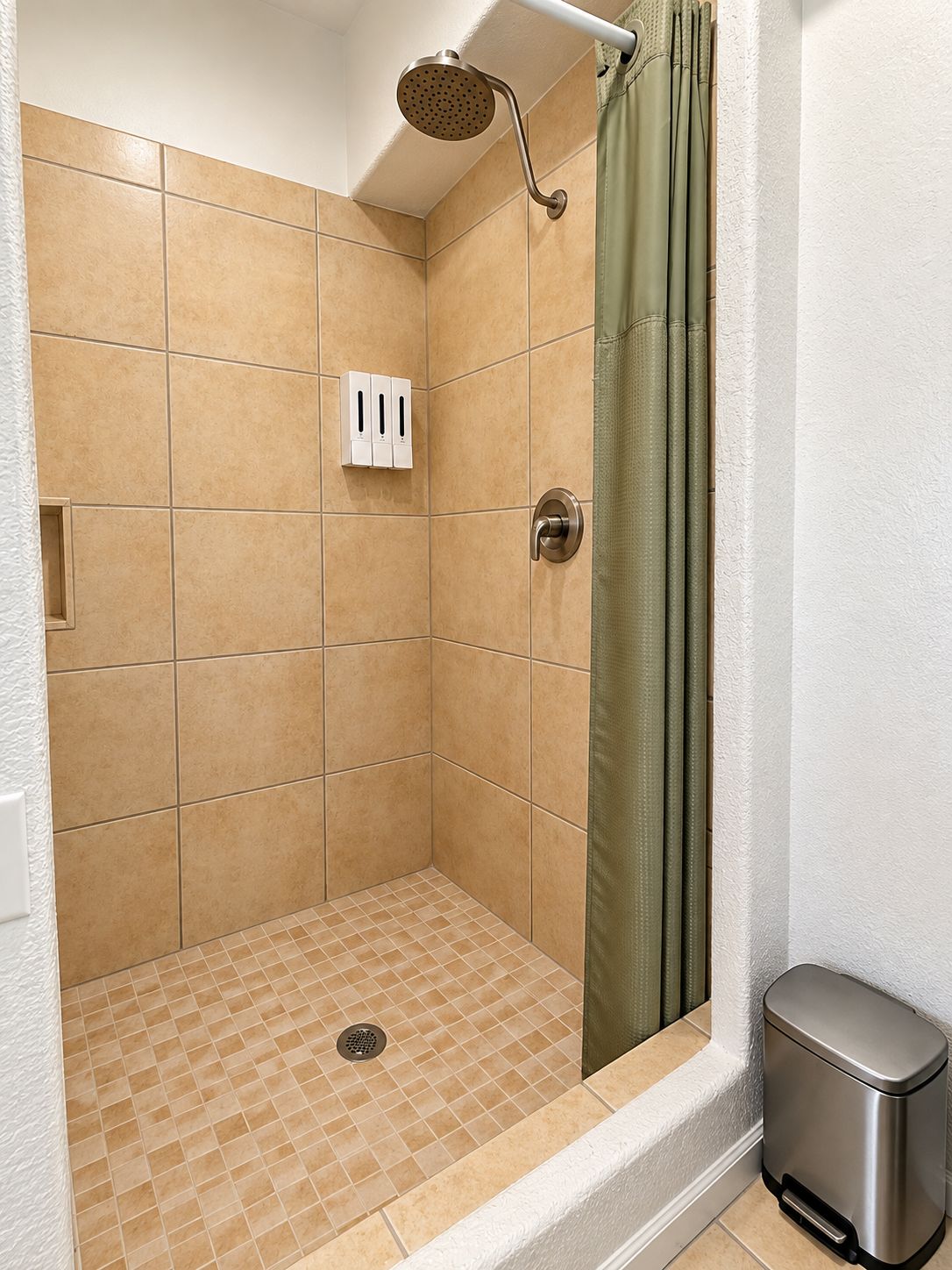 A shower area with a handheld showerhead, a green shower curtain, and a waste bin.