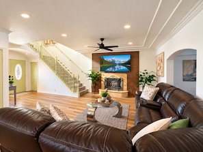 A spacious living room with a brown leather sofa, a coffee table, and a television mounted on a wooden wall. A staircase with a green railing leads to another room. - Fun For Everyone!  Family Friendly Pool Home! (Thousand Palms)