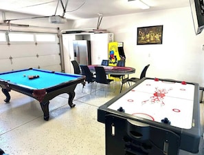 Een biljart, een tafelvoetbaltafel en een pingpongtafel in een ruimte met stoelen en een deur.