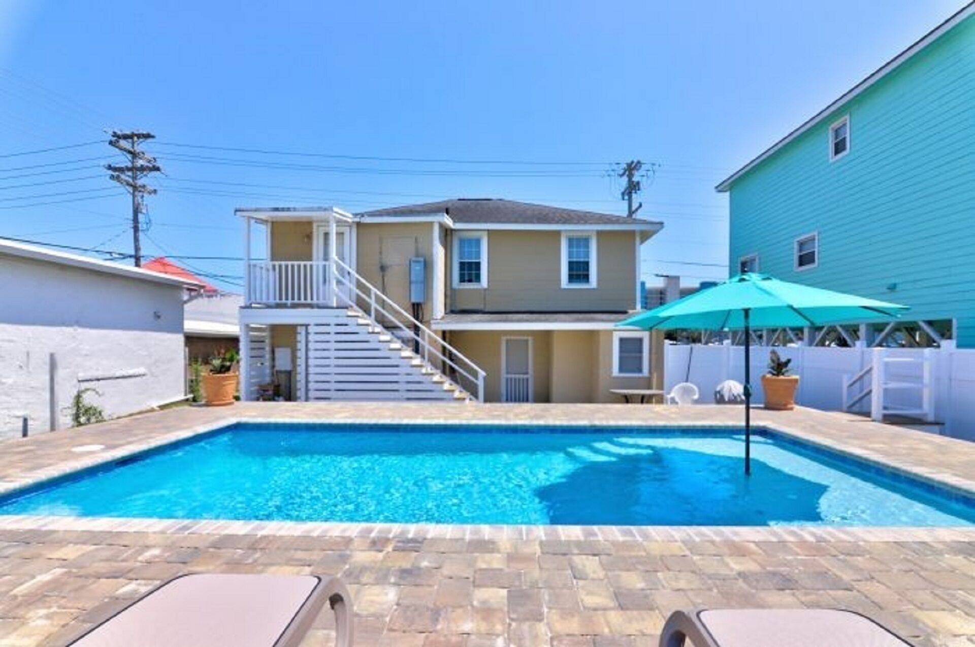 Una piscina residencial con una casa de dos pisos al fondo, un área de patio techada y un cielo azul despejado.