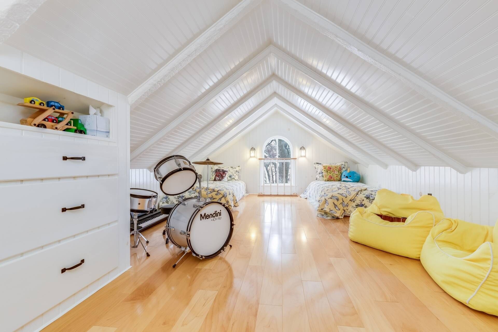 A spacious attic room with a high ceiling, wooden flooring, a drum set, a piano, and a cozy seating area with a yellow bean bag.