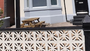 Uma casa de dois andares com um parapeito saliente, uma porta da frente preta e um pequeno jardim frontal com um banco de madeira.