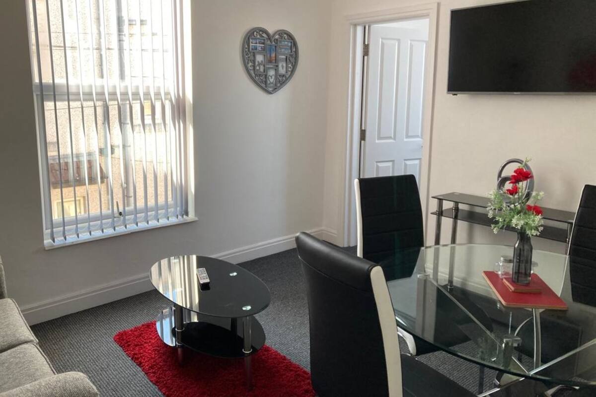 Un salon moderne avec une table à manger en verre, des fauteuils en cuir noir, un tapis rouge, un élément décoratif mural en forme de cœur et un téléviseur fixé au mur.