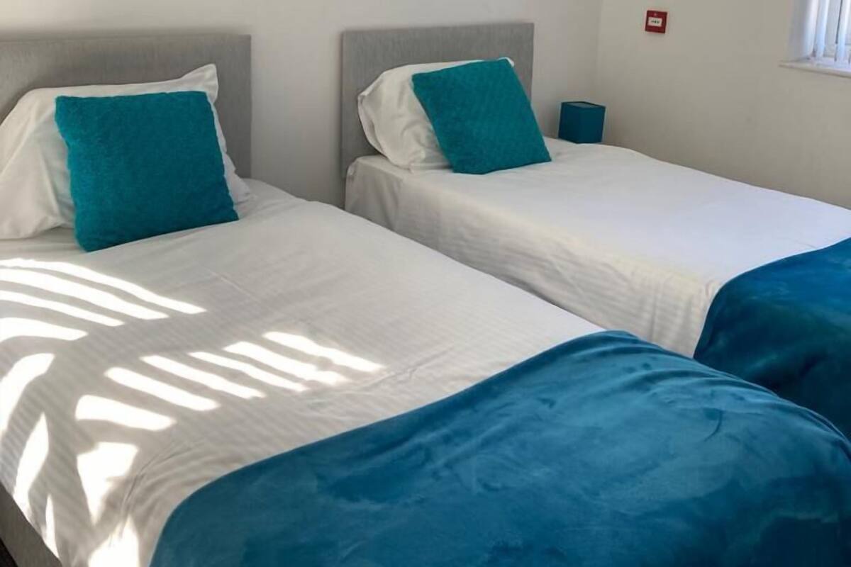 Ein Zimmer mit zwei Einzelbetten, jeweils mit grauem Kopfteil und einem türkisfarbenen Kissen. Rechts befindet sich ein Fenster und an der Wand eine kleine rote Notleuchte.
