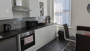 Cozinha moderna com armários brancos, bancada preta, micro-ondas e fogão. Há uma janela com persianas, uma cadeira e uma mesa.