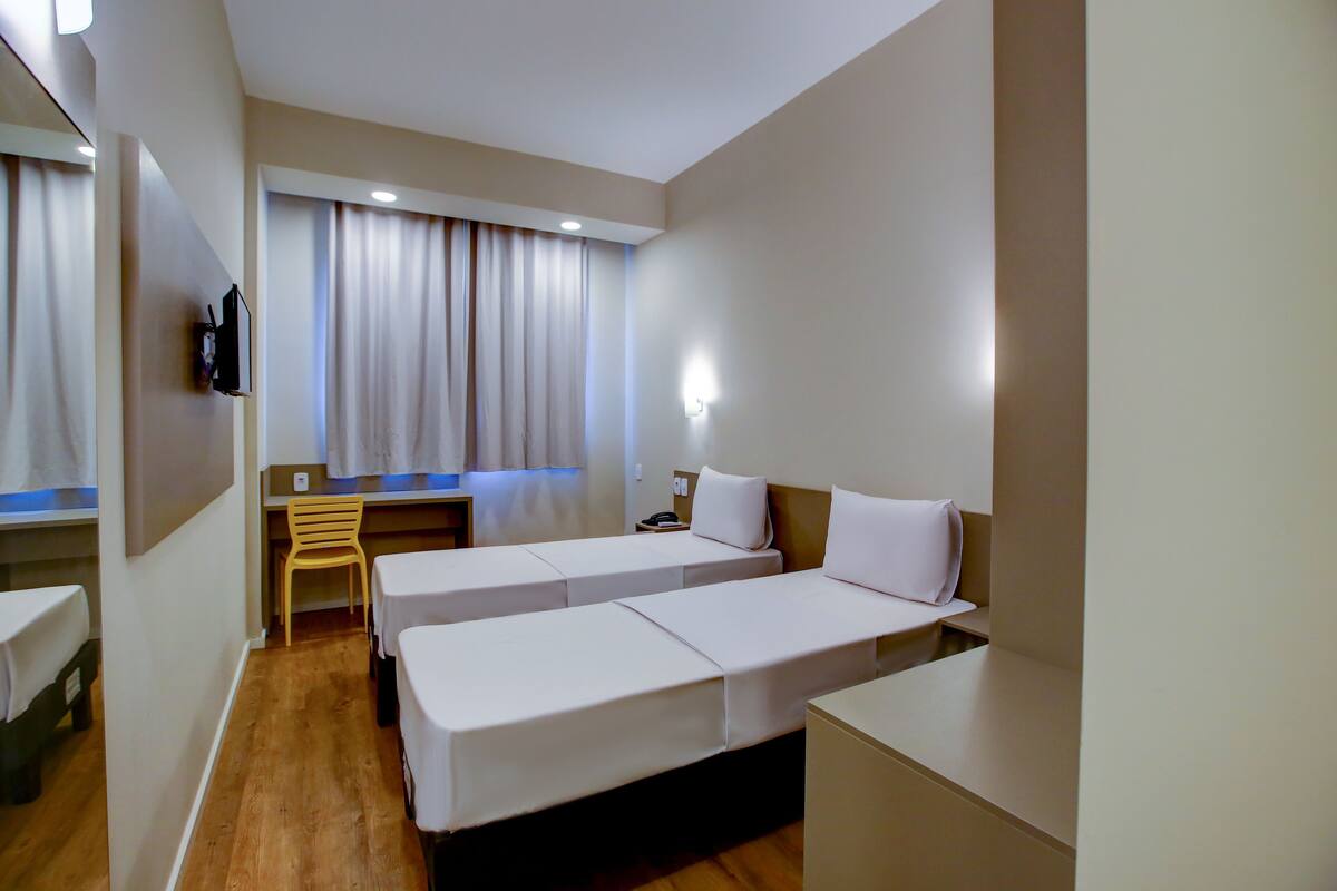 Ett hotellrum med två separata sängar, ett skrivbord, en stol, en TV och ett fönster med gardiner.