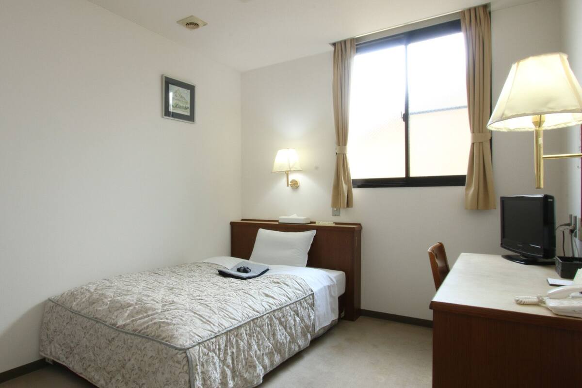 Habitación de hotel con una cama individual, un escritorio con televisor, una lámpara y una ventana con cortinas.