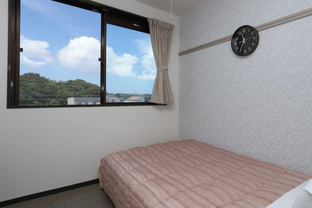 A bedroom with a bed, a window with curtains, a wall clock, and a patterned wallpaper.