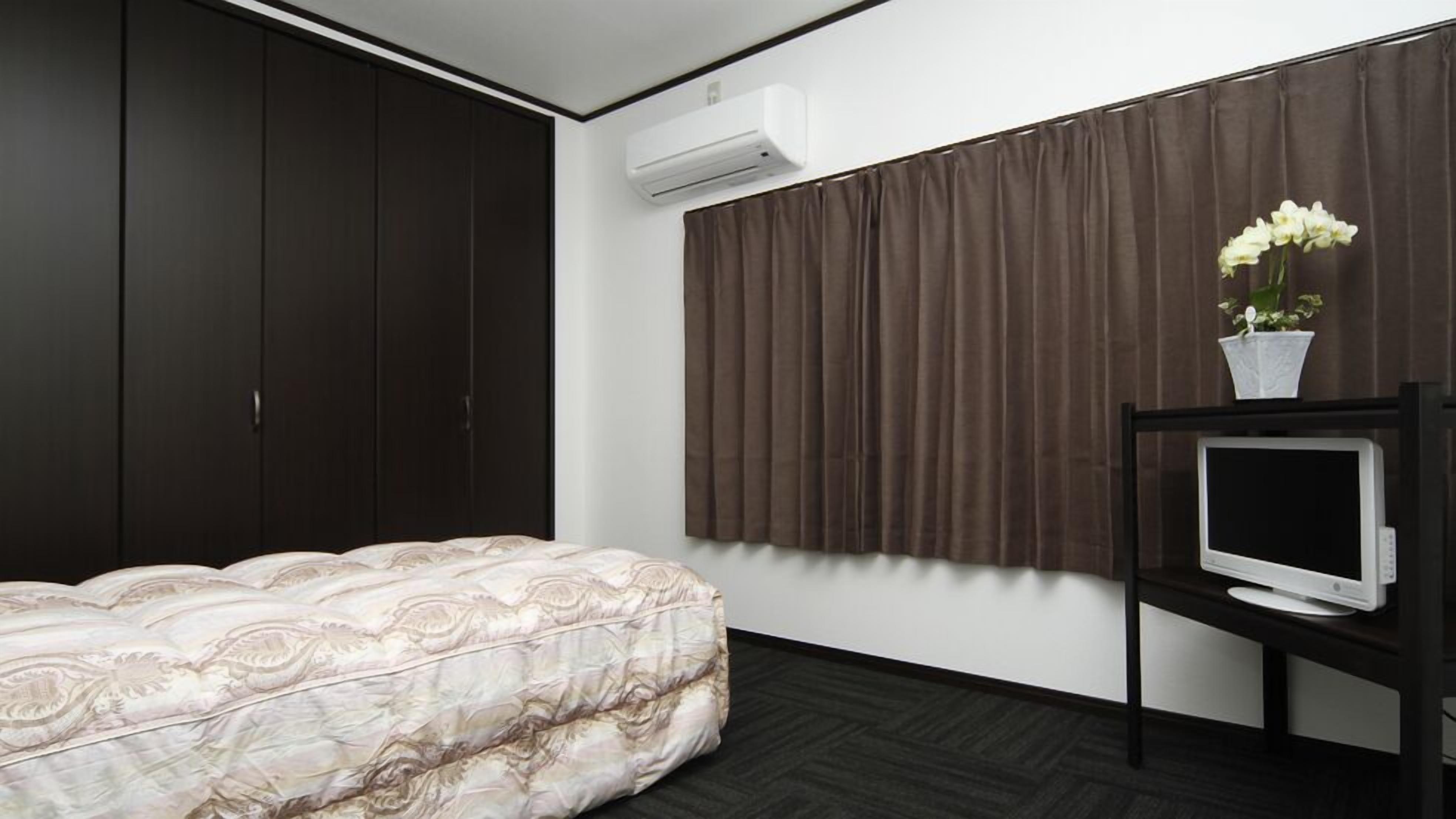 Une chambre à coucher comprenant un lit, une armoire sombre, un appareil de climatisation, des rideaux, une petite table avec un téléviseur et une plante en pot.
