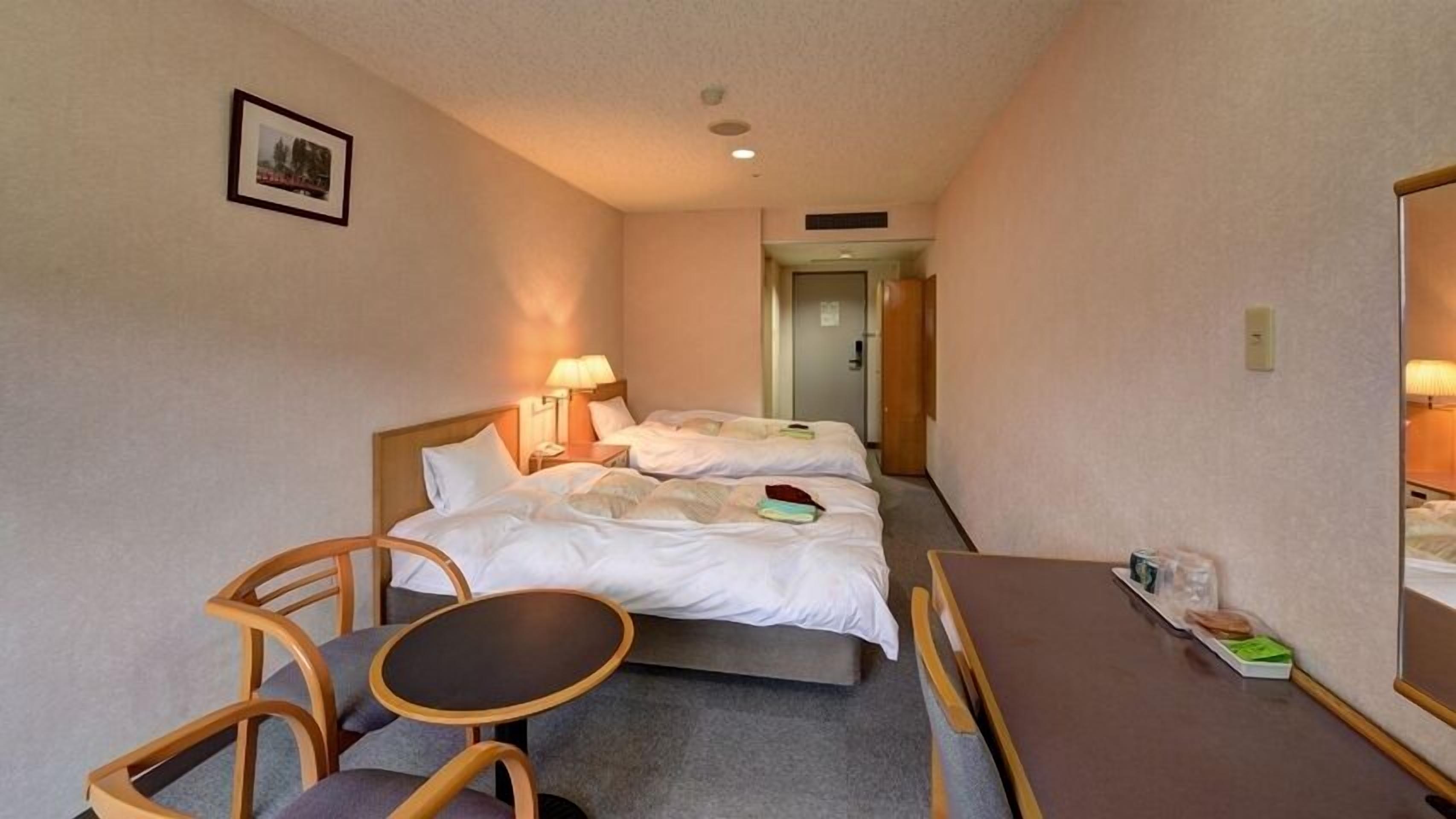 Una habitación de hotel con dos camas, una mesita, una silla y una lámpara de noche.