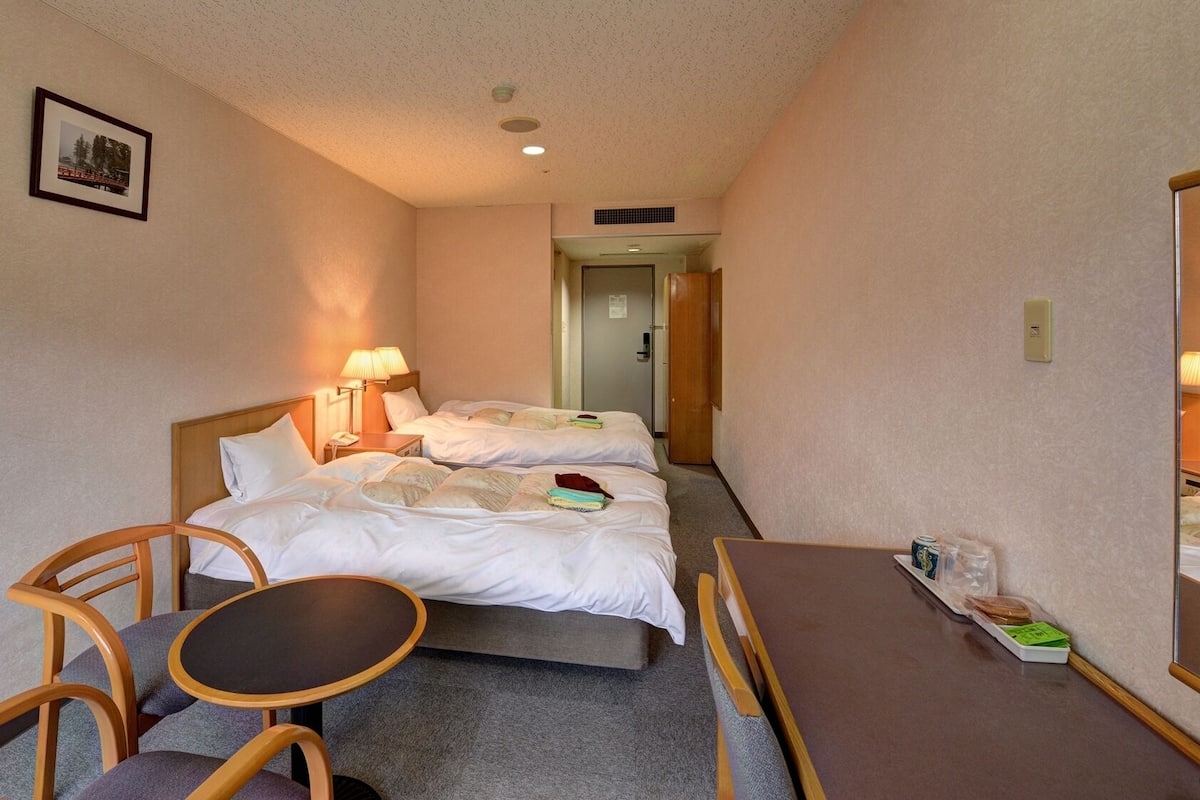 Hotellihuone, jossa on kaksi sänkyä, pieni pöytä, tuoli ja peili.