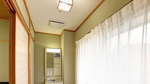 Una habitación pequeña con una silla de madera, un escritorio blanco, un espejo y una ventana con cortinas translúcidas.