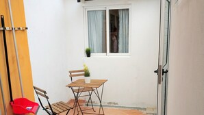 A small patio area with a wooden table and chairs, a potted plant, and a window with curtains.
