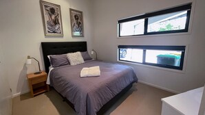 Une chambre à coucher comprenant un lit, des tables de chevet, une lampe, deux œuvres d’art encadrées et des fenêtres donnant sur l’extérieur.