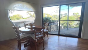 Un coin repas avec une table ronde et des chaises, une grande fenêtre et une vue sur une terrasse et la verdure environnante.