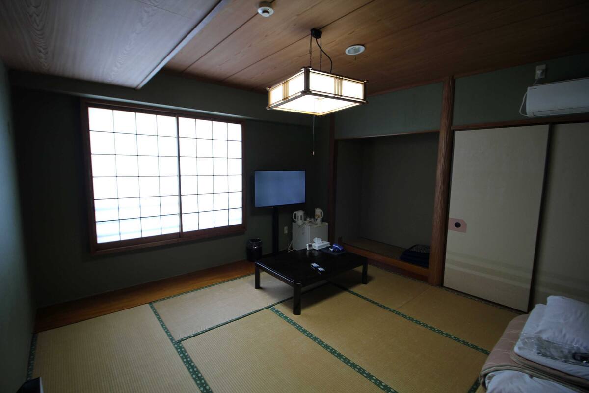 Ein traditioneller japanischer Raum mit Tatami-Bodenbelag, einem niedrigen Tisch, einem Fernseher und gleitenden Türen.