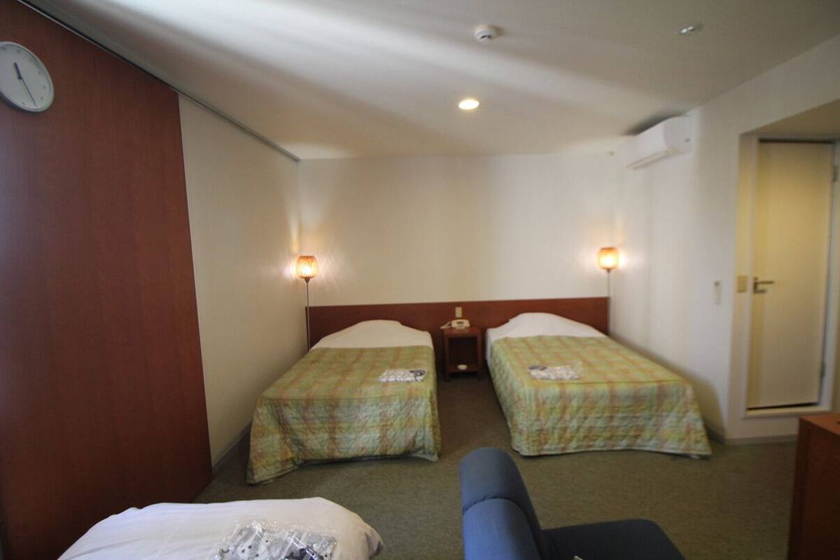 Ein Hotelzimmer mit zwei Einzelbetten, einem kleinen Tisch, einem Stuhl, einer Wanduhr und zwei Wandleuchten.