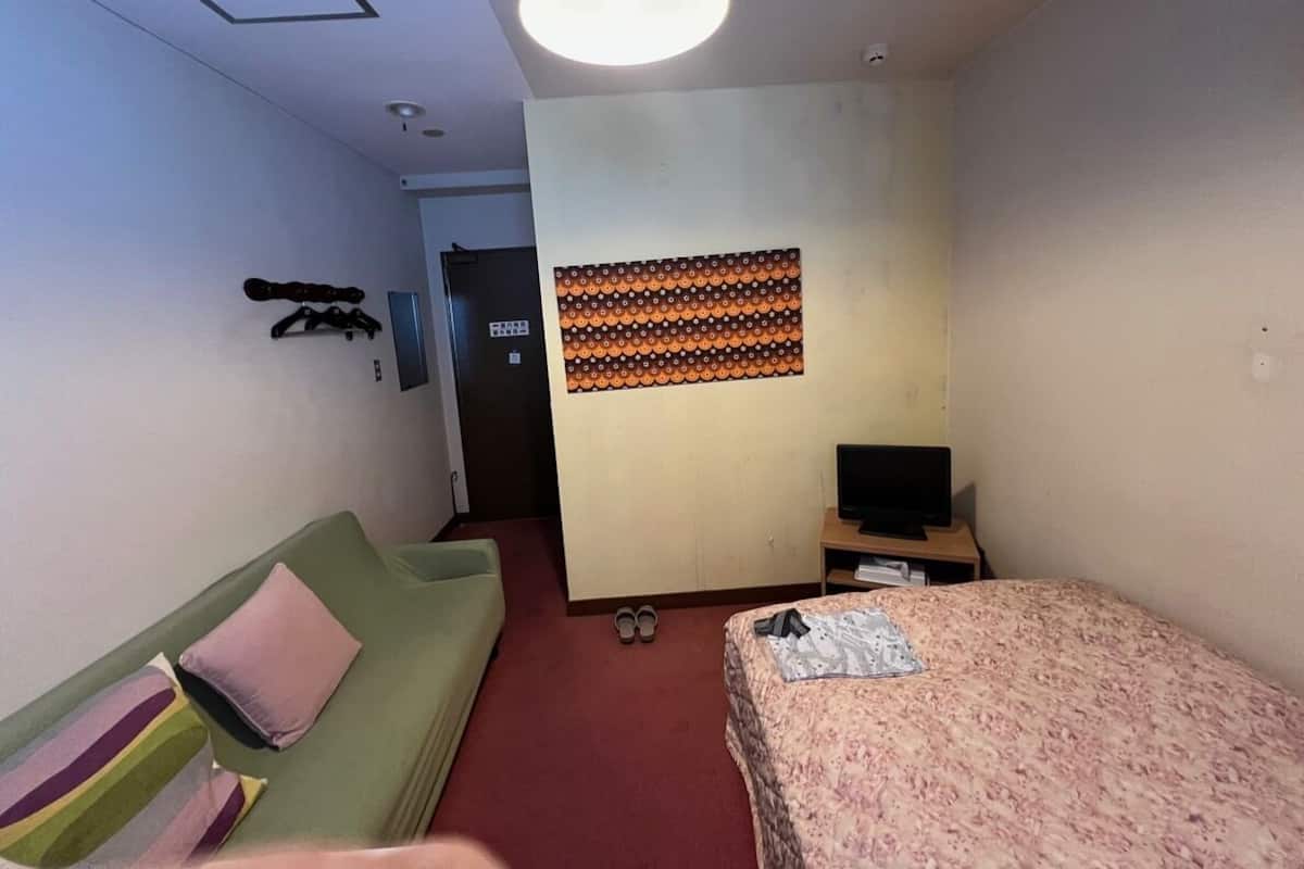 A hotel room with a green sofa, a bed with a floral bedspread, a television on a wooden stand, and a patterned wall hanging.