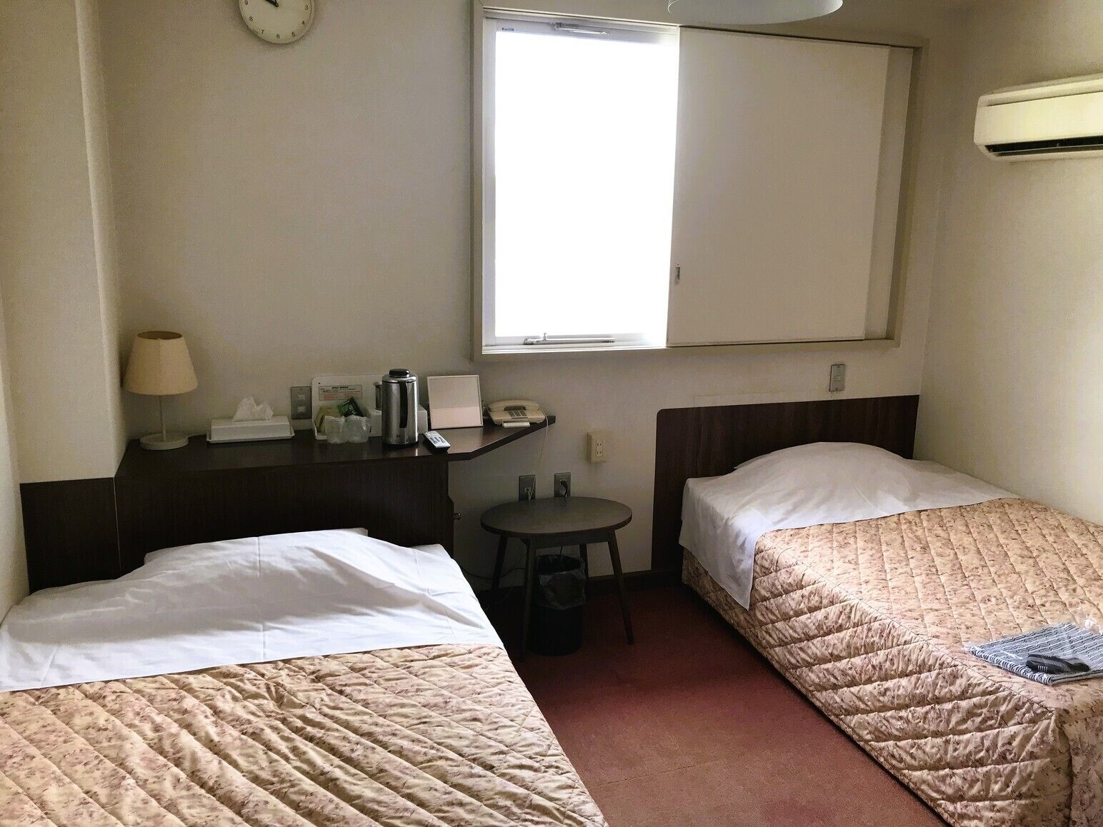 Ein Hotelzimmer mit zwei Betten, einem Schreibtisch mit Lampe, einem kleinen Tisch, einem Mülleimer und einem Fenster.