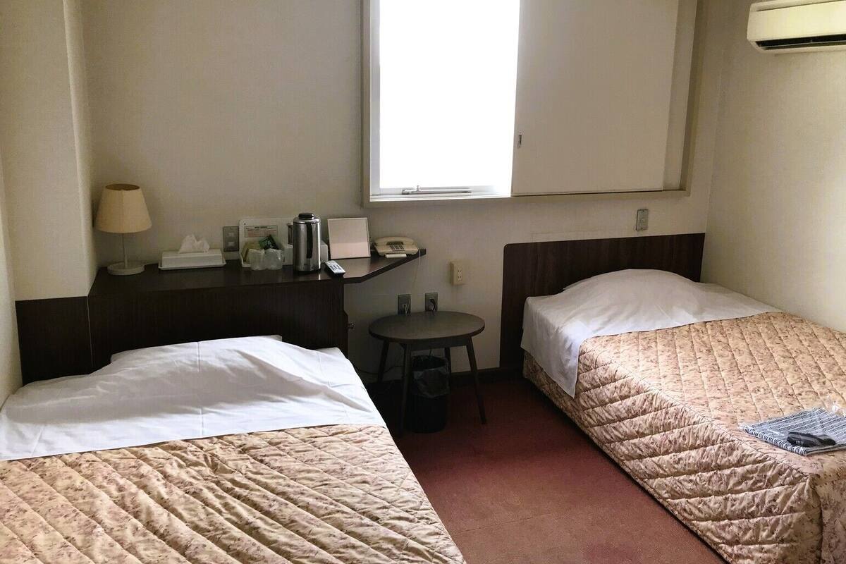 Ein Hotelzimmer mit zwei Betten, einem Schreibtisch mit Lampe, einem kleinen Tisch, einem Mülleimer und einem Fenster.