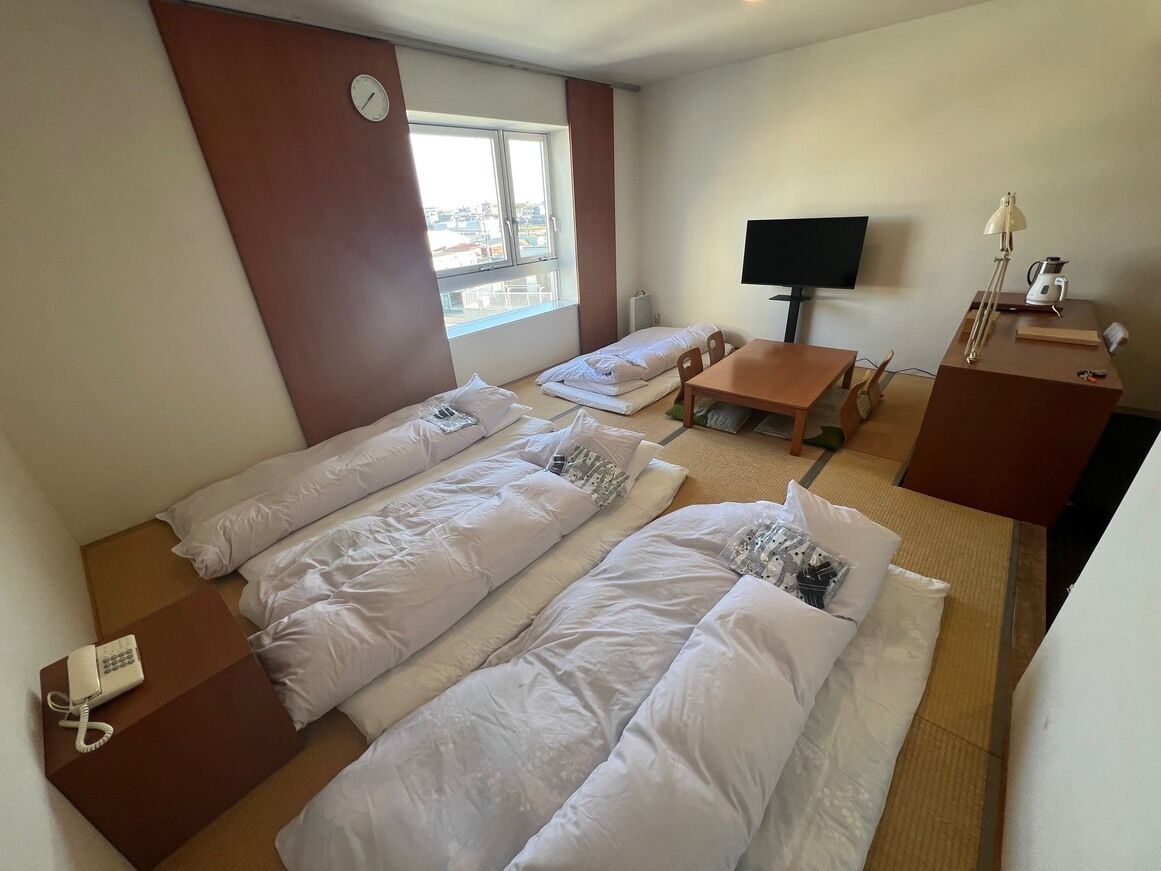 Ein Hotelzimmer mit drei Betten, einem Fernseher, einem Schreibtisch mit Lampe und einem Fenster.