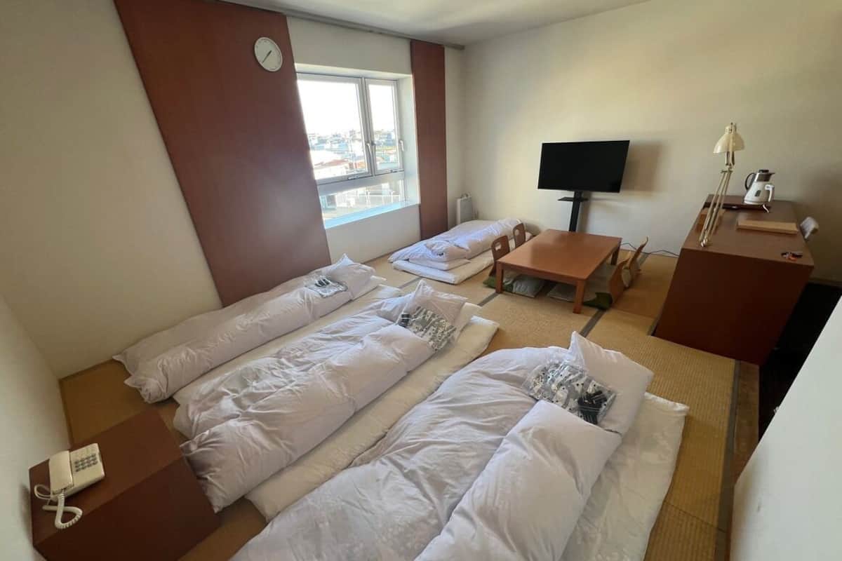 Ein Hotelzimmer mit drei Betten, einem Fernseher, einem Schreibtisch mit Lampe und einem Fenster.