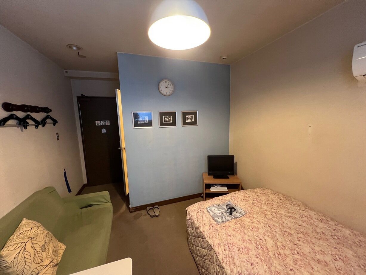 Ein Zimmer mit einem Bett, einem grünen Schlafsofa, einem Fernseher auf einem Holztischchen, einer Uhr an der Wand und einer Tür.