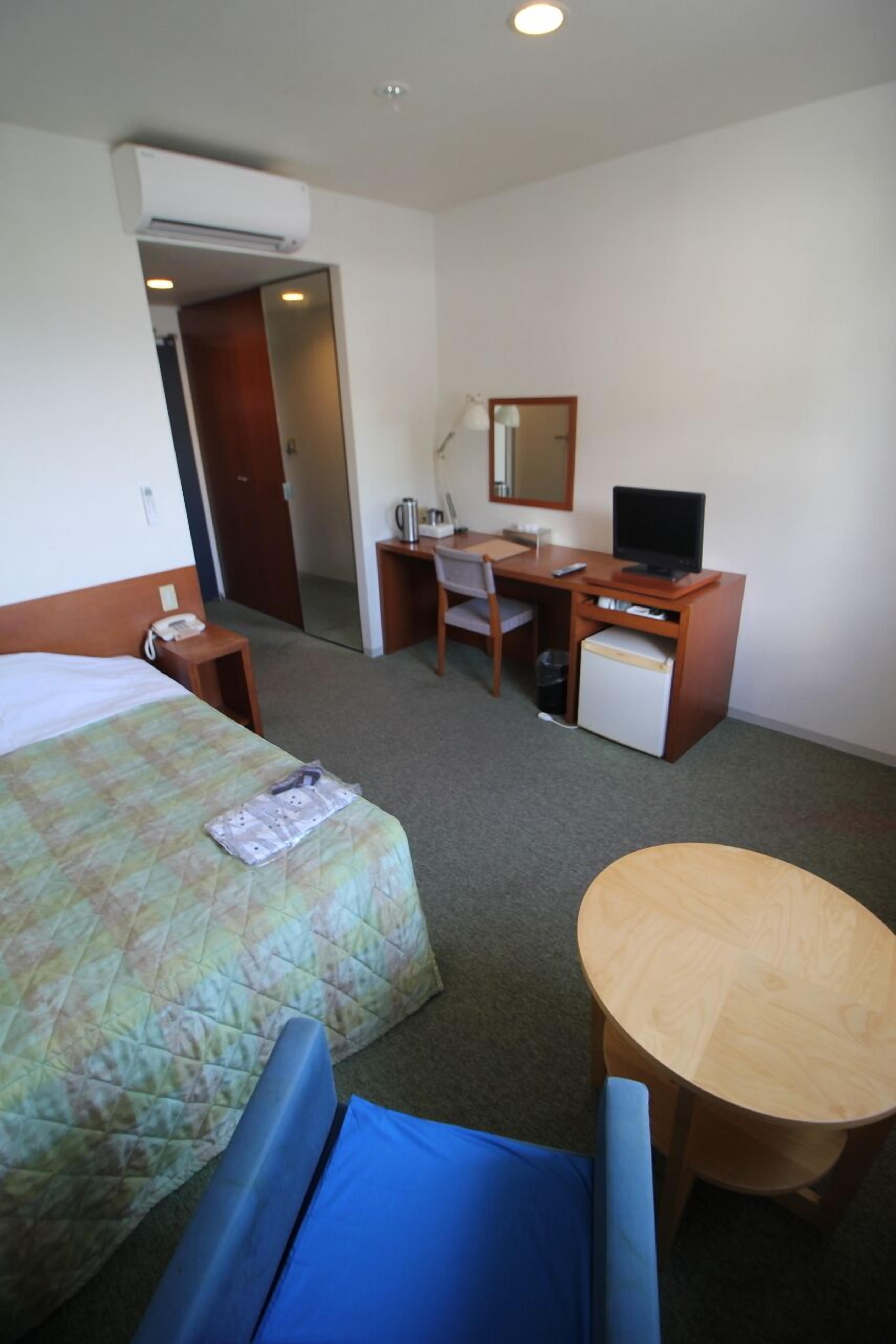 Ein Hotelzimmer mit Bett, Schreibtisch, Stuhl, kleinem Kühlschrank und einem runden Tisch.