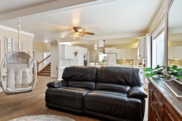 A living room with a black leather sofa, a wicker chair, a ceiling fan, and a view into the kitchen.