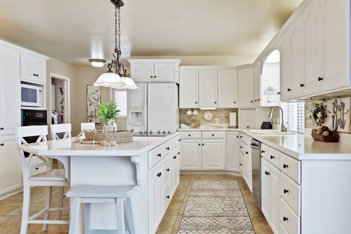 A kitchen with white cabinetry, a central island with bar stools, a hanging light fixture, and a patterned rug.