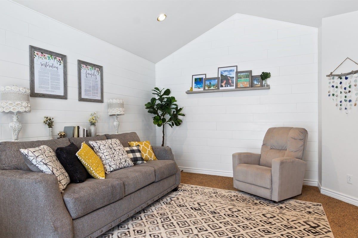 Een woonkamer met een grijze bank, een kamerkleed met dessin, en een stoel. Er hangen ingelijste foto's aan de muur, een plant en een plank met decoratieve objecten.