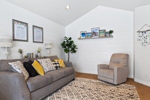 A living room with a grey sofa, a patterned rug, and a chair. There are framed pictures on the wall, a plant, and a shelf with decorative items.