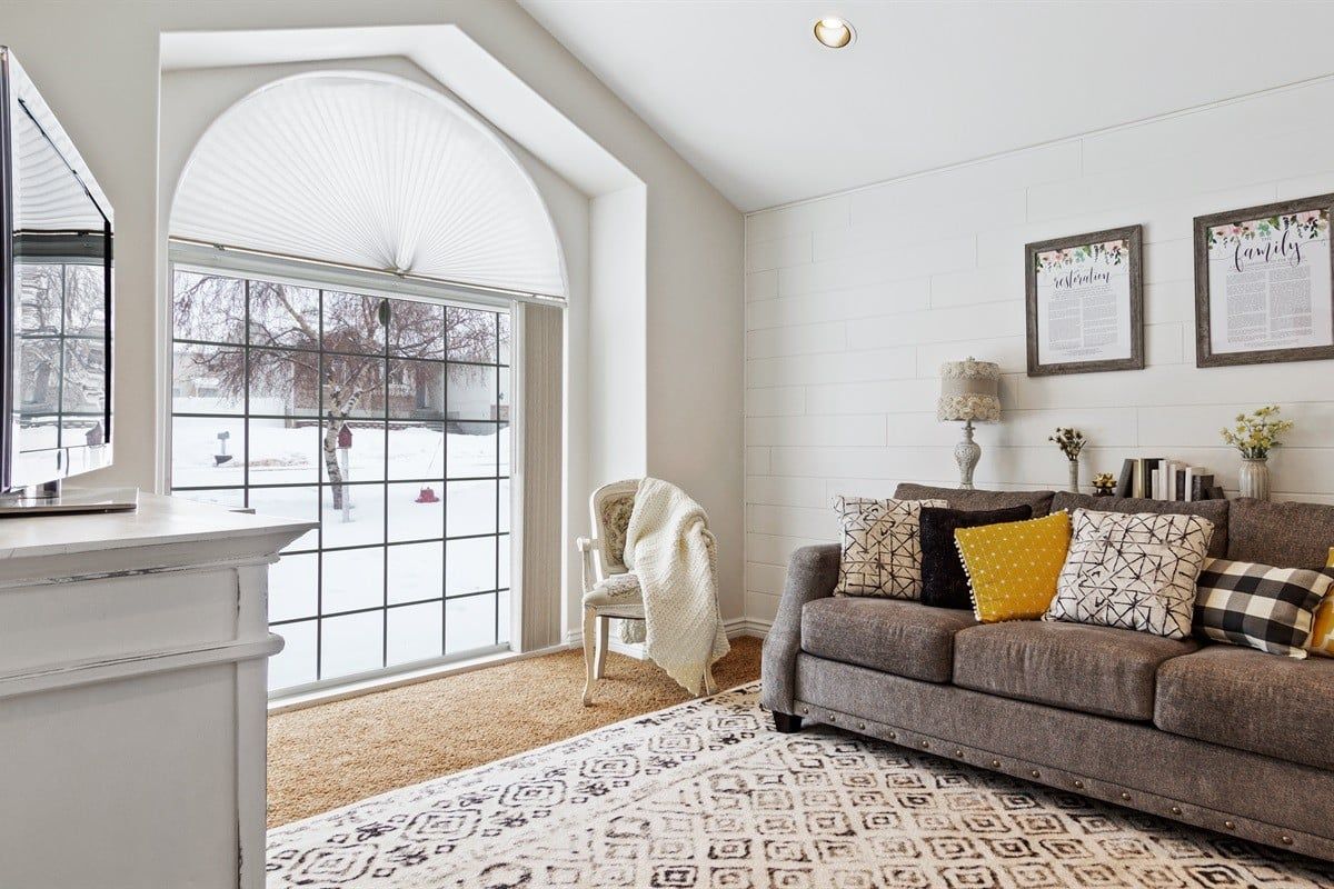 A living room with a large window, a sofa with patterned cushions, and a white wall with framed text.