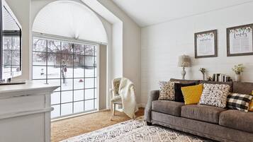 A living room with a large window, a sofa with patterned cushions, and a white wall with framed text.