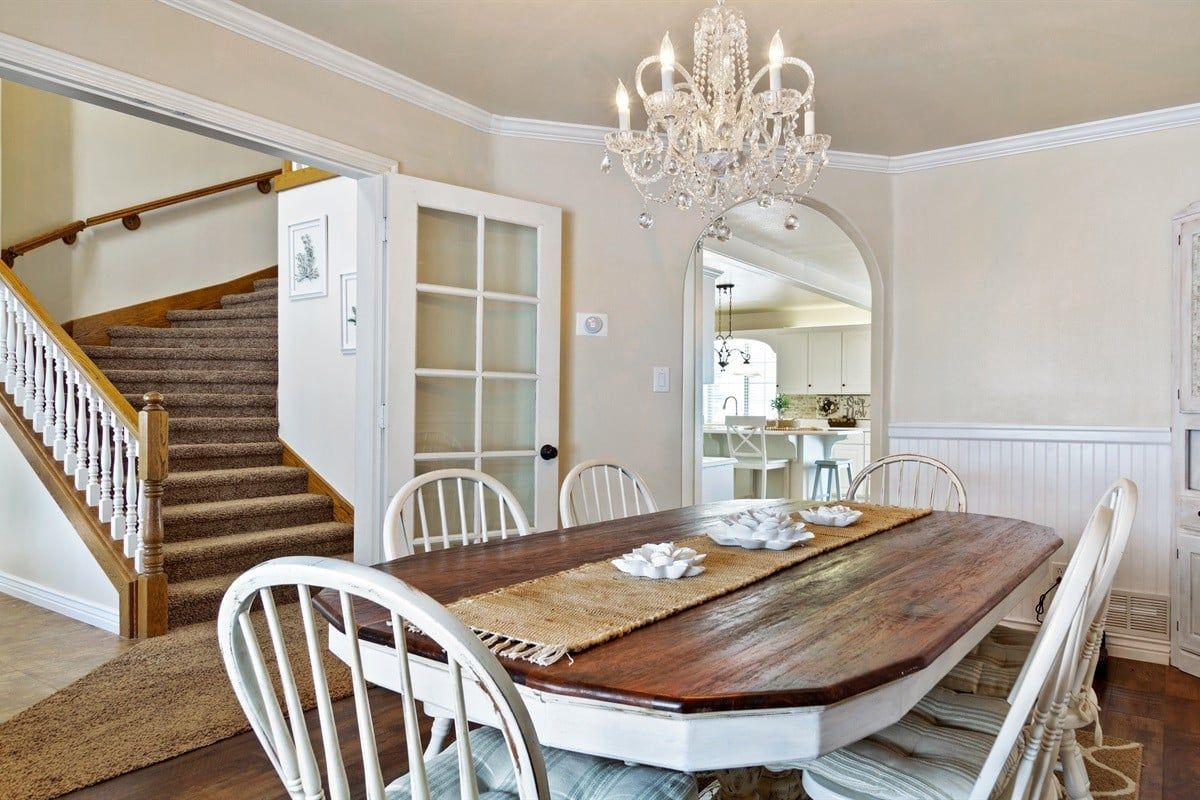 A dining area with a wooden table, white chairs, a chandelier, and a staircase.