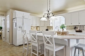 A kitchen with white cabinets, a central island with chairs, a refrigerator, and a hanging light fixture.