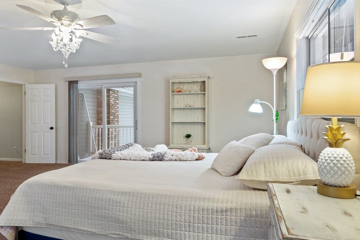 A bedroom with a large bed, a ceiling fan, a nightstand with a lamp, and a window with blinds.