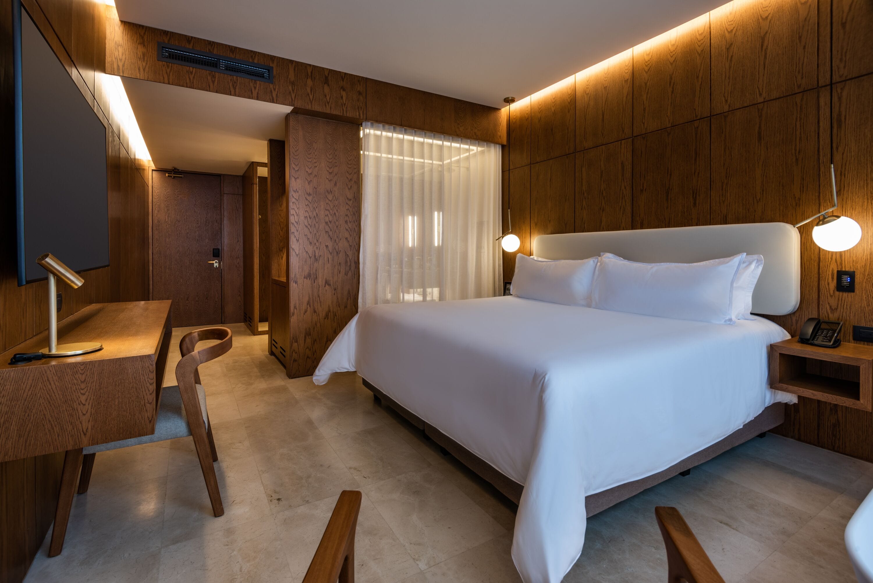 A modern hotel room with a large bed, wooden panel walls, a desk with a chair, and a bedside table with a lamp.