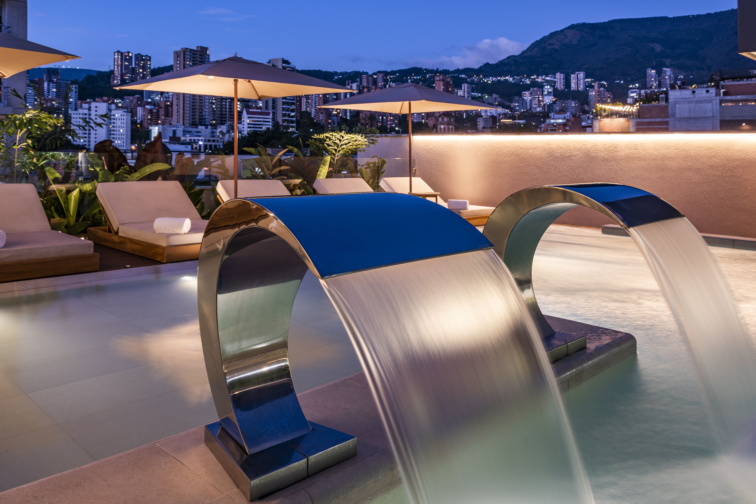 Un espace piscine aménagé sur le toit, avec des chaises longues et des parasols, offrant une vue sur le paysage urbain, avec ses bâtiments et une colline.
