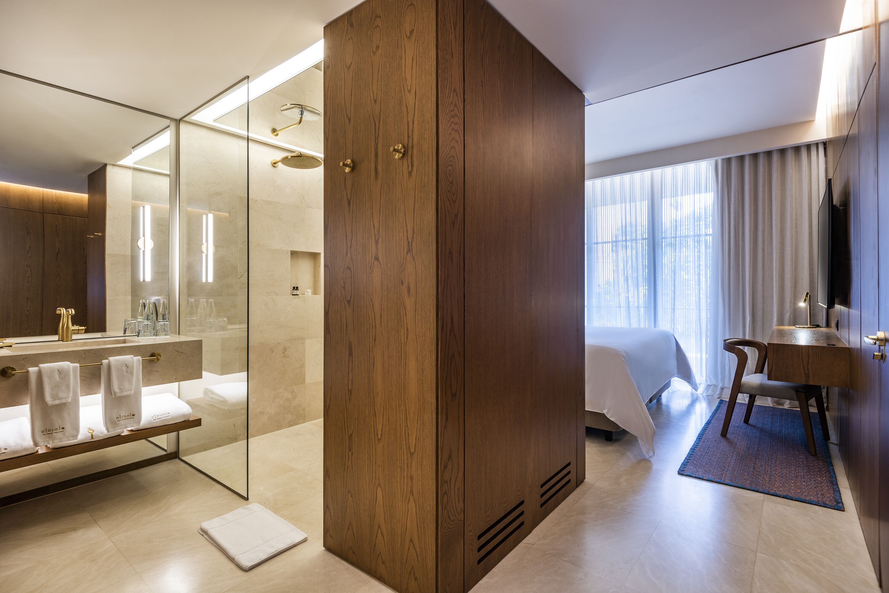 A modern hotel room with a glass-enclosed shower, a wooden wardrobe, a bed with a white bedspread, a desk with a chair, and a large window with curtains.