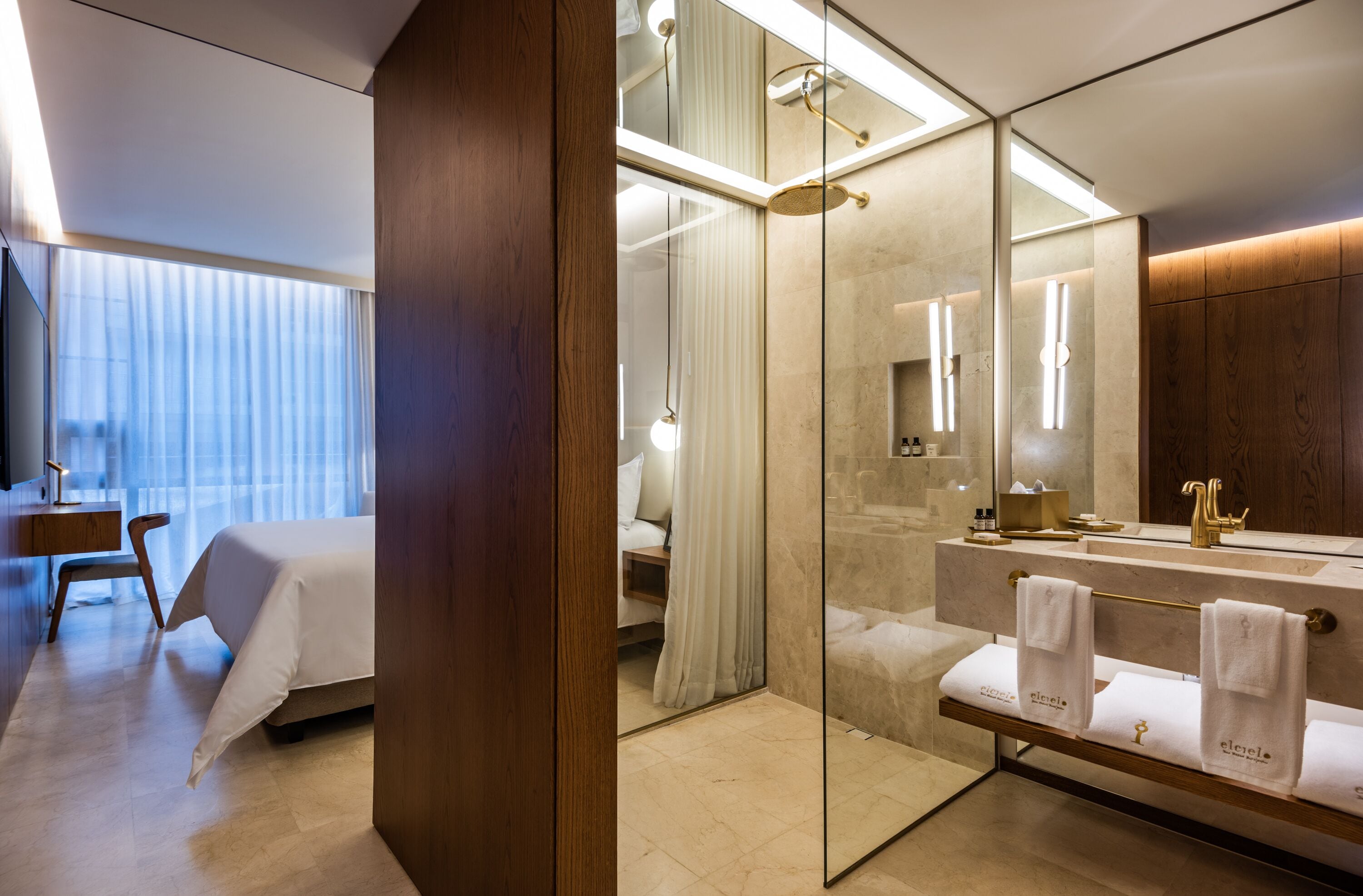 Una habitación de hotel moderna con una cama grande, un escritorio de madera, una cabina de ducha de vidrio y un tocador con espejo y toallero.