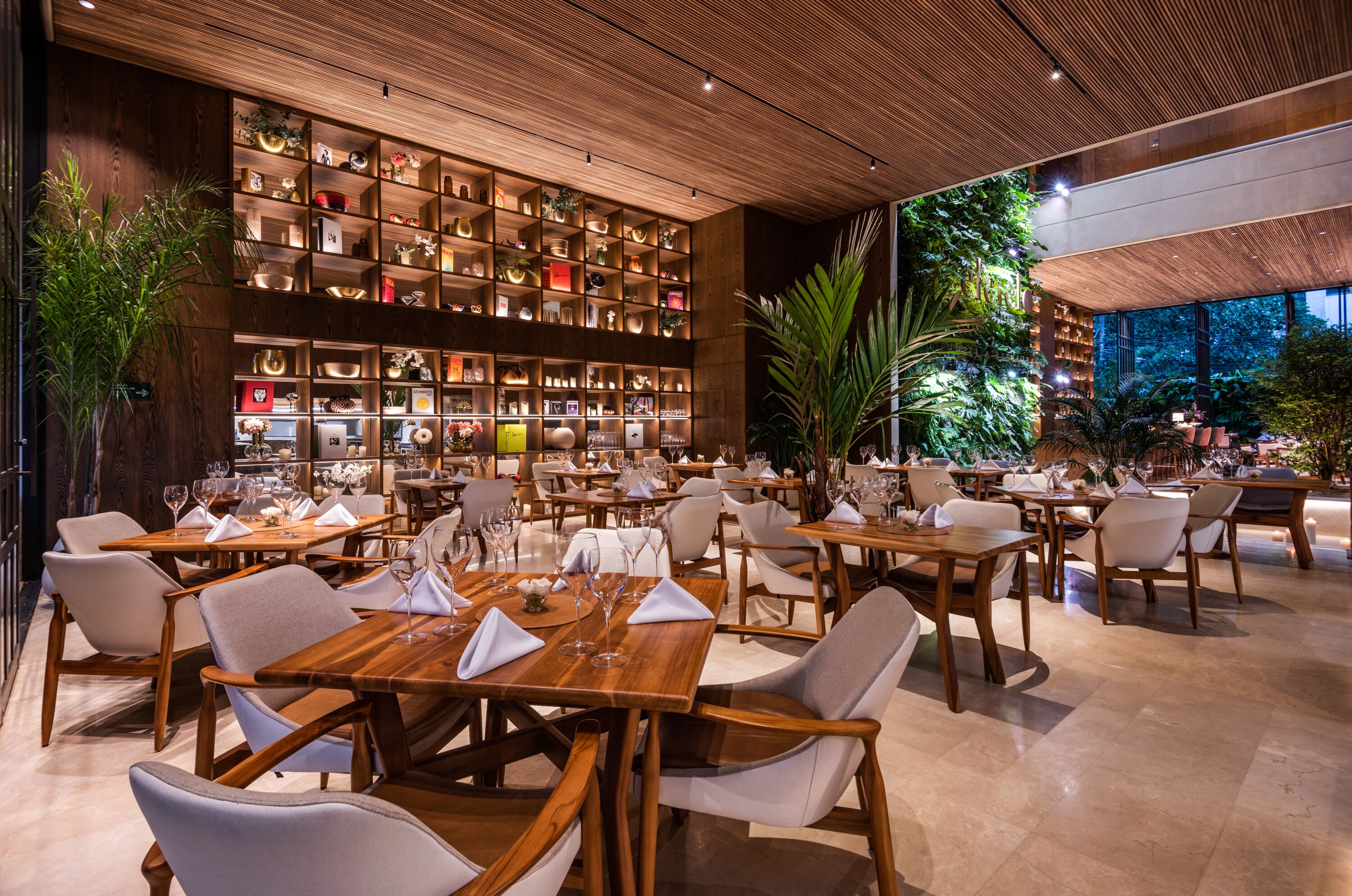 A spacious restaurant with wooden tables and chairs, a large bookshelf filled with books and decorative items, and a view of the outdoors.
