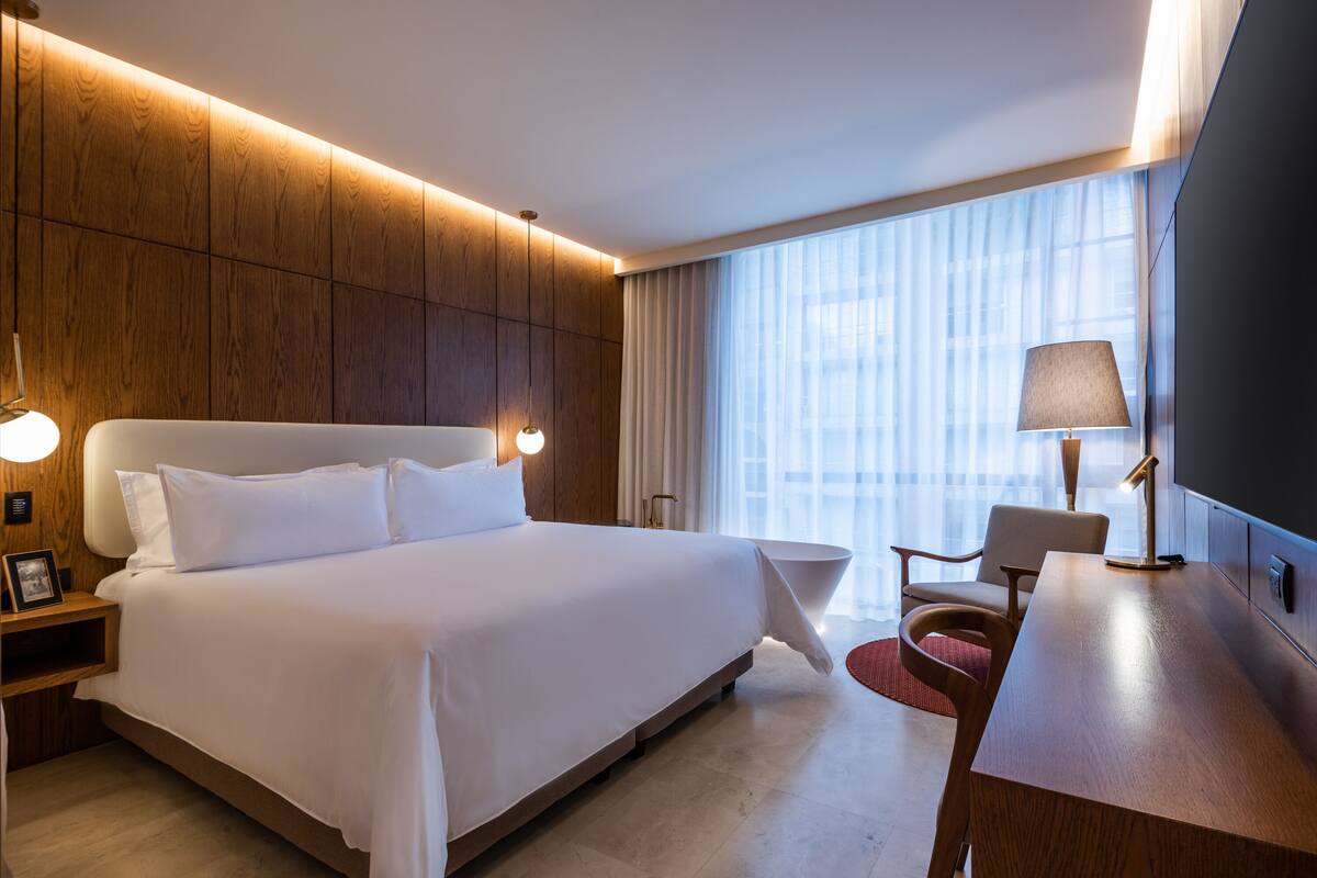 Une chambre d’hôtel moderne dotée d’un grand lit, de lampes de chevet, d’un bureau et offrant une vue sur la ville grâce à de grandes fenêtres.