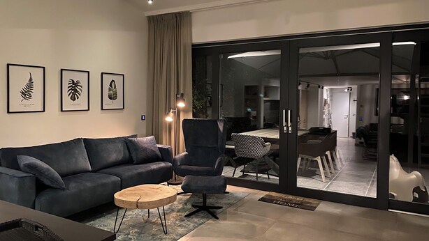 Moderni olohuone, jossa on tummanharmaa sohva, pyöreä sohvapöytä ja kuviollinen matto. Seinällä on kehystettyjä kasvitaiuluja, ja huoneesta on liukuovien kautta pääsy ruokailutilaan. Lisäksi huoneessa on pieni sivupöytä, jonka päällä on tarjotinalusta.
