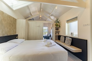 A bedroom with a skylight, a bed with white linens, a bedside table with a radio, and a view of the outdoors.