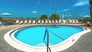 A swimming pool with lounge chairs and umbrellas, surrounded by a wooden deck and palm trees, with a beach view in the background.